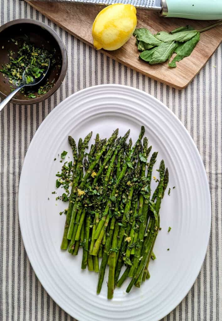 Overhead tablescape shot of a platter of roasted asparagus topped with herbs and lemon zest on a white platter with a brown cutting board off center. The cutting board has a microplane with a lemon plus mint leaves. Off center is a small brown bowl with the mint gremolata.