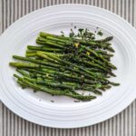 Overhead shot of fresh, green roasted asparagus topped with mint gremolata on a white platter. The platter sits on a tray stripped tablecloth.