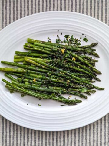 Overhead shot of fresh, green roasted asparagus topped with mint gremolata on a white platter. The platter sits on a tray stripped tablecloth.
