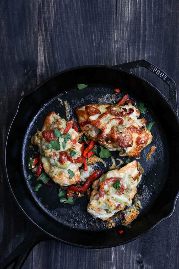 An overhead shot of a cast iron skillet with cooked chicken covered with artichokes, roasted red peppers and smothered in melted cheese. The skillet is on a rustic black table.