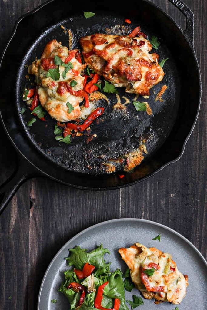 An overhead shot of a cast iron skillet with chicken covered in artichoke hearts, roasted red peppers and melted jack cheese. Off center is a gray plate with a single serving of the chicken and a side green salad.