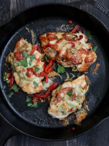 An overhead shot of a cast iron skillet on a rustic black table with cooked chicken topped with artichokes and roasted red peppers and melted cheese.