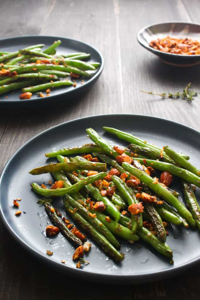 Finished serving of seared green beans with toasted spiced almonds on round gray plate. In the background is a second plate of finished green beans and a bowl of extra spiced almonds.