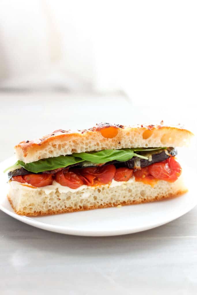 A closeup of a single serving of a roasted eggplant and tomato sandwich on focaccia with fresh basil sitting on a round white plate.