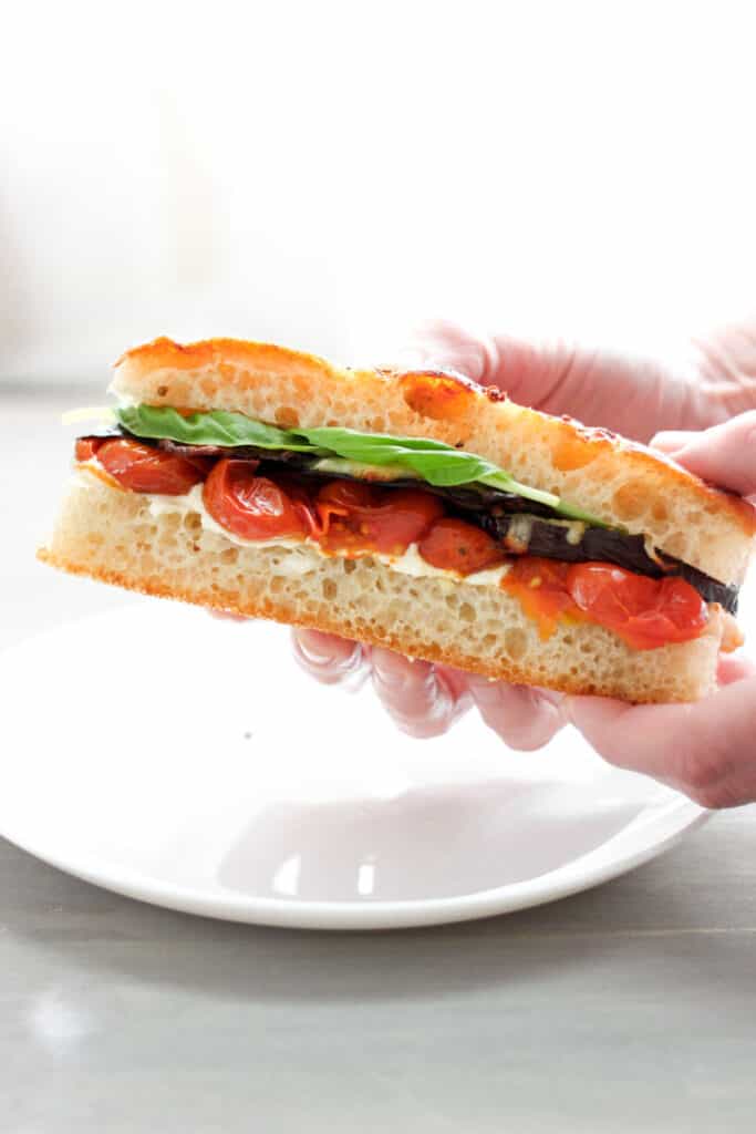 Two hands holding a serving of a roasted eggplant and tomato sandwich on focaccia with fresh basil. The hands are holding the sandwich just off a round white plate.