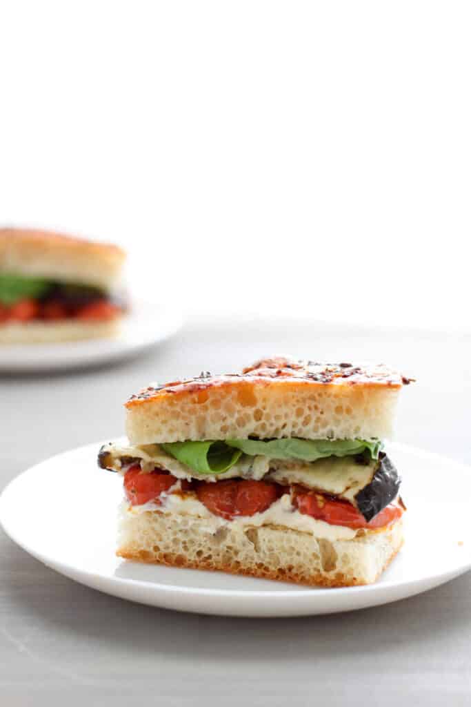 Single portion of a roasted eggplant and tomato sandwich on focaccia bread with fresh basil sitting on a round white plate with another serving on a white plate blurred in the background.