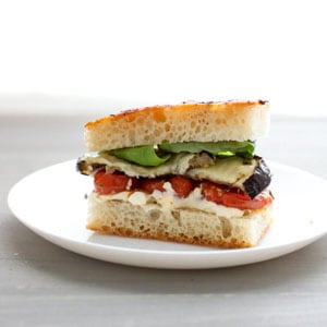 Single serving of a roasted eggplant and tomato sandwich on focaccia bread with basil sitting on a round white plate with a blurry white background.