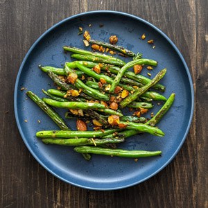Seared green beans on a blue plate topped with toasted spiced almonds on a gray countertop.