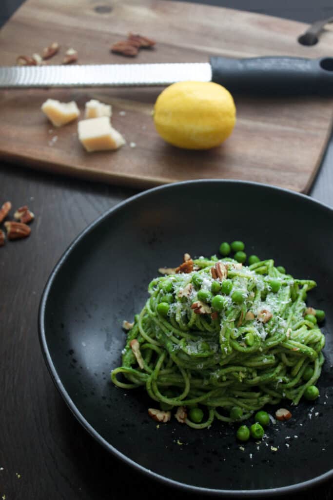 A single portion of creamy spinach pasta on a round black bowl topped with grated parmesan cheese, chopped toasted pecans and green peas. In the background out of focus is a cutting board with a microplane grater, a lemon and parmesan cheese.