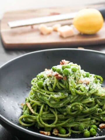 A single portion of creamy spinach pasta on a round black bowl topped with grated parmesan, toasted chopped pecans and green peas. Out of focus is a cutting board with a micrpplane grater, a lemon and parmesan cheese.