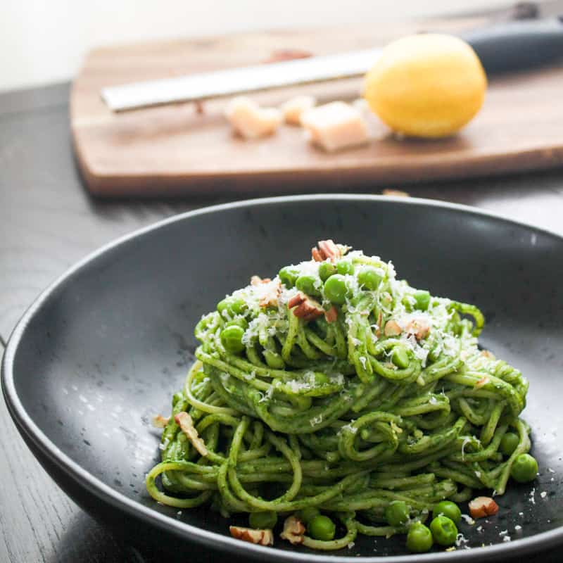 A single portion of creamy spinach pasta on a round black bowl topped with grated parmesan, toasted chopped pecans and green peas. Out of focus is a cutting board with a micrpplane grater, a lemon and parmesan cheese.