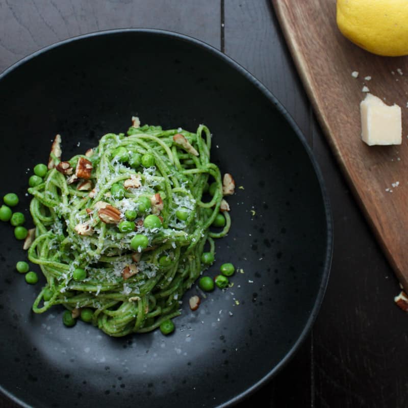 An overhead shot of a single portion of creamy spinach pasta in a black bowl topped with grated parmesan cheese, chopped toasted pecans and green peas. Just to the side is a cutting board with a lemon and parmesan cheese.