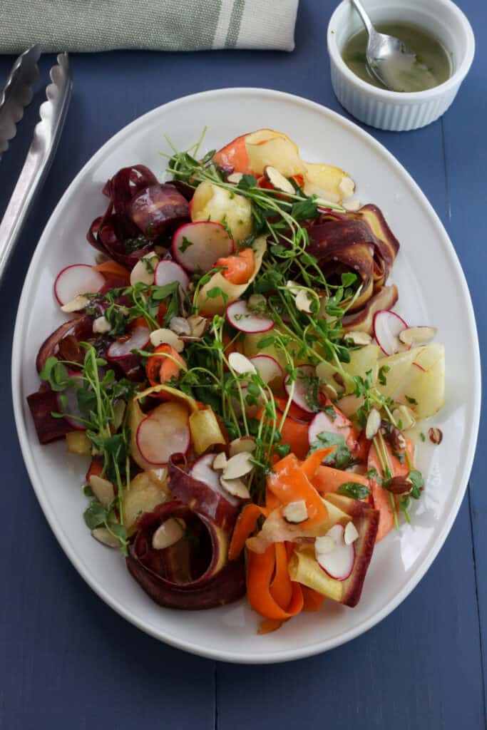 A vibrantly colorful platter of thinly sliced yellow, orange and purple carrots with green pea shoots, sliced almonds and thinly sliced radishes on a white platter. Slightly off center is a small white bowl with a lemon and herb dressing. The background table is blue.