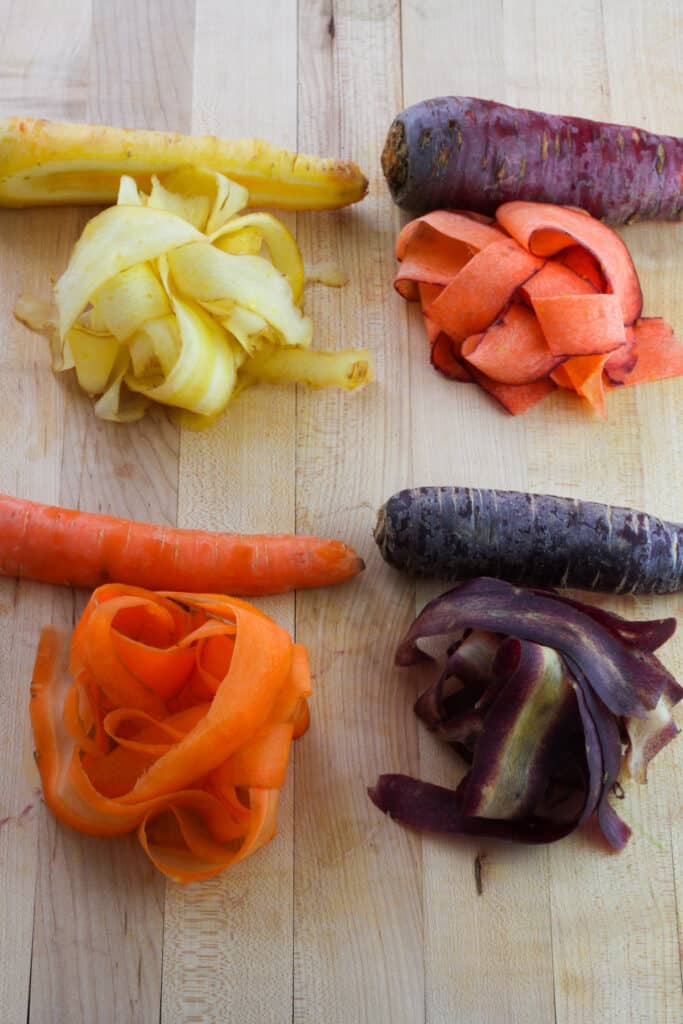 An overhead shot on a butcher block cutting board of a yellow, purple, red and orange carrot with piles of thinly sliced carrot in front of each carrot.