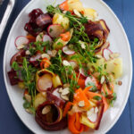 A salad on white platter on a blue table with ribbons of multicolored carrots, green pea shoots, thinly sliced radishes and sliced almonds dressed with lemon and herb vinaigrette.