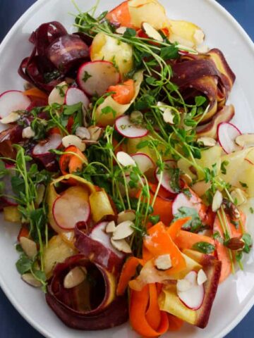 A salad on white platter on a blue table with ribbons of multicolored carrots, green pea shoots, thinly sliced radishes and sliced almonds dressed with lemon and herb vinaigrette.