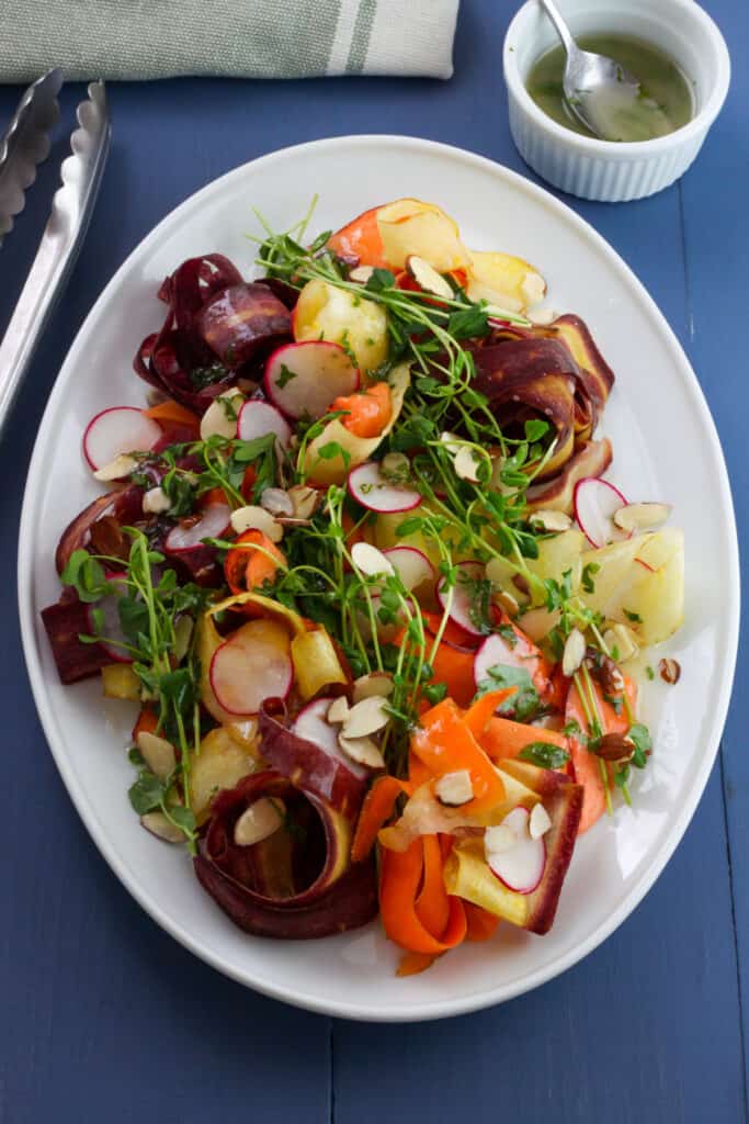 An Overhead shot of a white platter with multicolored carrot ribbons, green pea shoots, thinly sliced radishes, sliced almonds drizzled with a lemon and herb dressing.