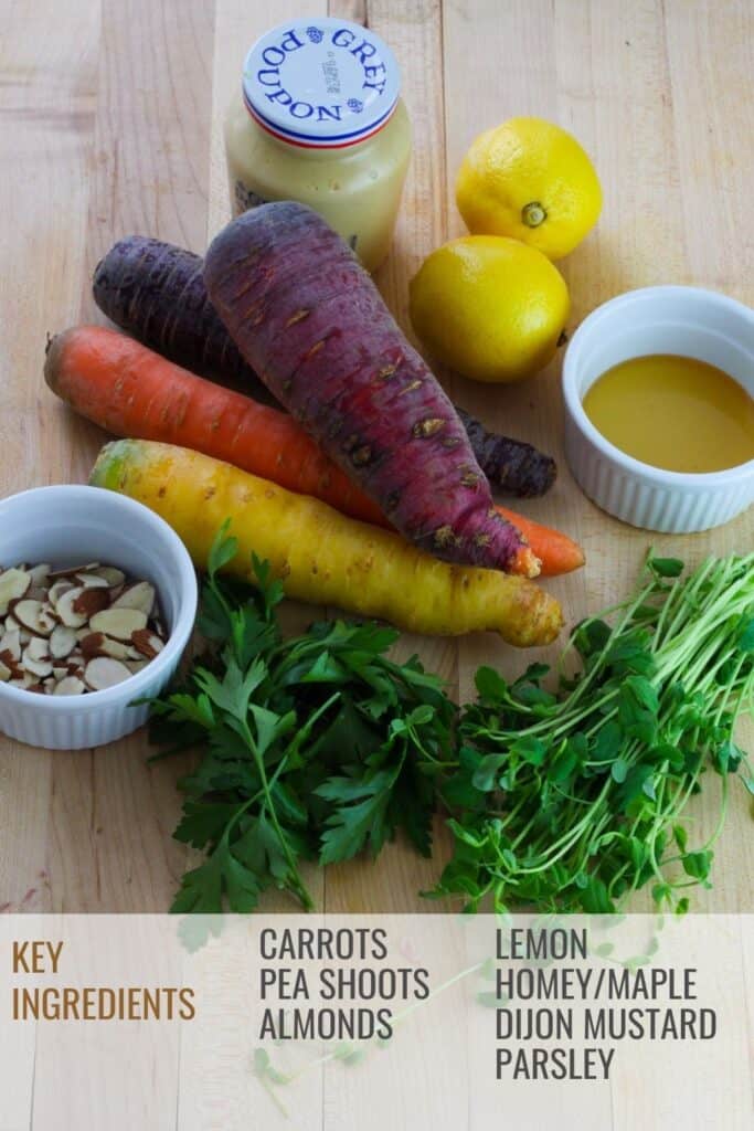 Overhead shot of multicolored whole carrots , lemons, mustard, honey, pea shoots, parsley and a small white bowl with sliced almonds all on a butcher block cutting board.