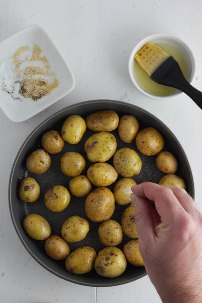 Overhead shot of a gray bowl with baby gold potatoes and a hand sprinkling salt and pepper on top with a small white bowl containing salt, pepper and spices and another bowl with oil and a pastry brush.