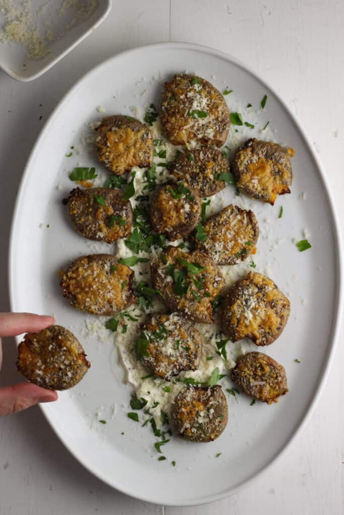 Overhead shot of a white platter with air fryer smashed potatoes on a mustard and mayonnaise sauce sprinkled with parsley and cheese and a hand just off center holding one potato.