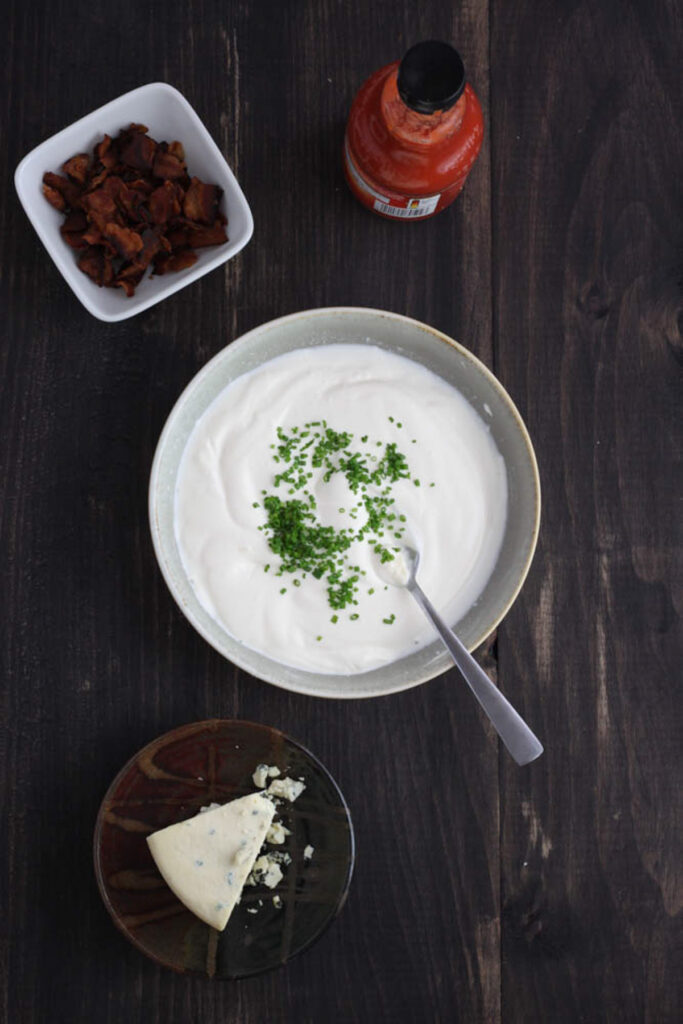 A gray bowl with mayonnaise, sour cream and lemon juice mixed together and chopped chives on top. Off center is a bowl of blue cheese, chopped bacon and a bottle of hot sauce.