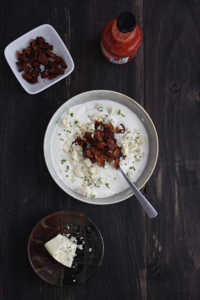 A gray bowl with mayonnaise, sour cream and lemon juice stirred together and chives and in the middle is chopped bacon and blue cheese unmixed. Off center is a bowl of chopped bacon, another bowl of blue cheese and a bottle of hot sauce.