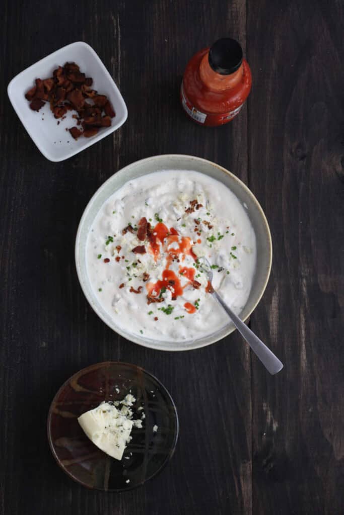 A gray bowl with mayonnaise, sour cream and lemon juice stirred together and chives and in the middle is chopped bacon and blue cheese and hot sauce. Off center is a bowl of chopped bacon, another bowl of blue cheese and a bottle of hot sauce.