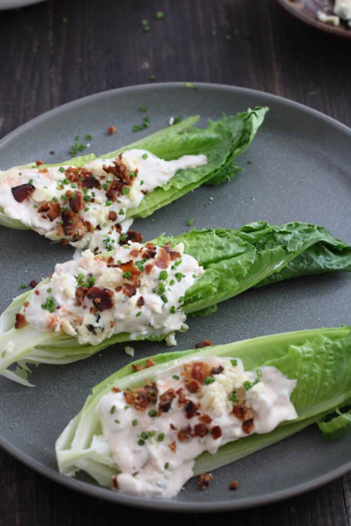 A blue plate with wedges of romaine lettuce topped with buffalo blue cheese dressing, chopped bacon and chives on a rustic black table.