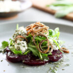 A close-up shot of sliced purple beets topped with arugula, goat cheese rolled in herbs, crispy fried shallots all drizzled with a lemon and herb vinaigrette on a gray plate.