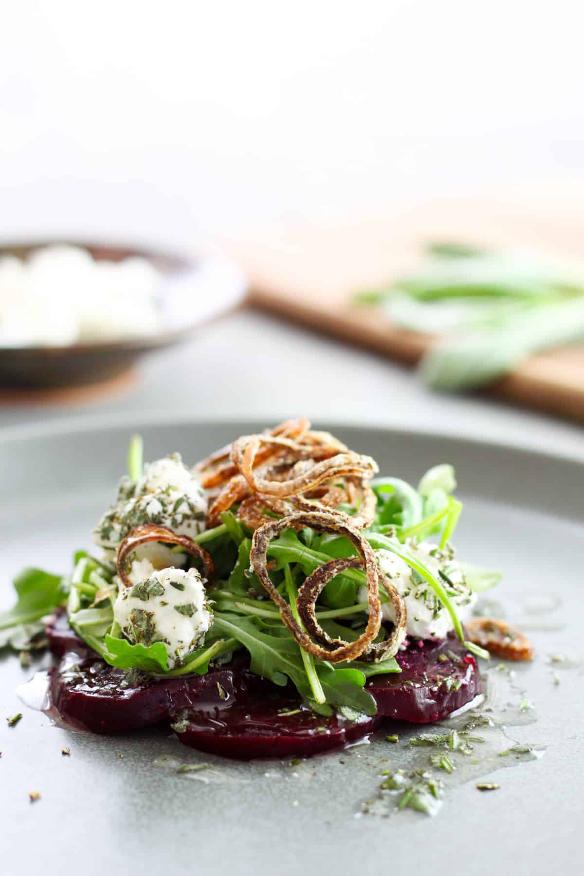A close up of sliced purple beets topped with arugula, goat cheese rolled in fresh herbs, and topped with crispy fried shallots all on a gray plate. Out of focus in the background is more goat cheese on a small bowl and herbs on a cutting board.