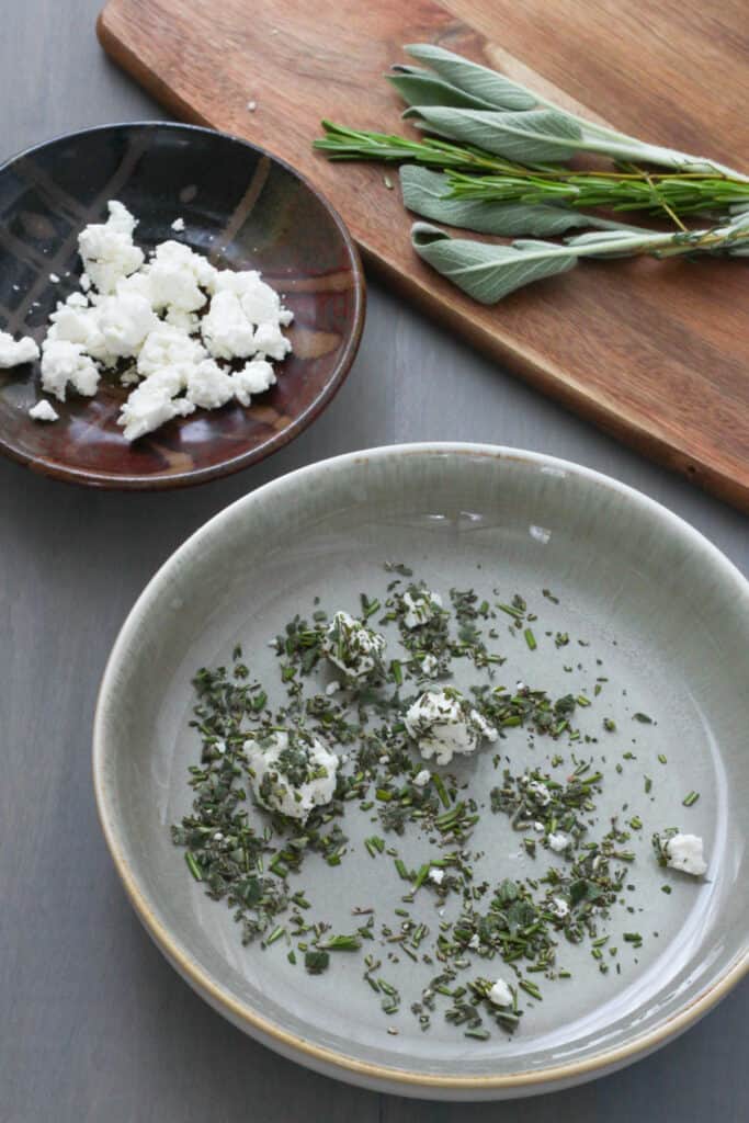 A gray bowl with chopped sage, rosemary and thyme with crumbled goat cheese coated in the herbs. Off center is a small brown bowl with crumble goat cheese and a brown cutting board with sage, rosemary and thyme.