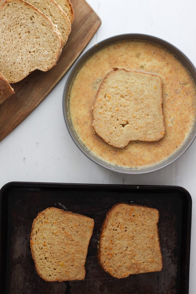 Sliced of bread dipping in a brown sugar and cinnamon custard. A sheet tray is just off center with pieces of bread already dipped resting plus a cutting board with slices of bread.