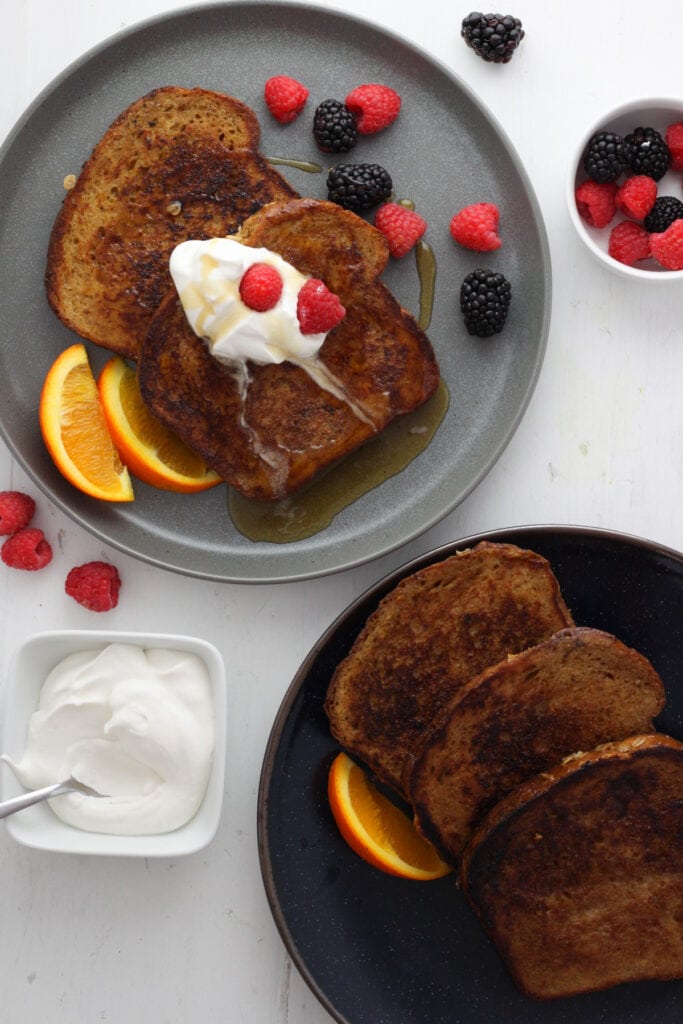 A gray plate with two slices of brown sugar french toast topped with whipped cream, berries and slices of orange. Off center is a platter of more french toast and a small bowl of whipped cream.