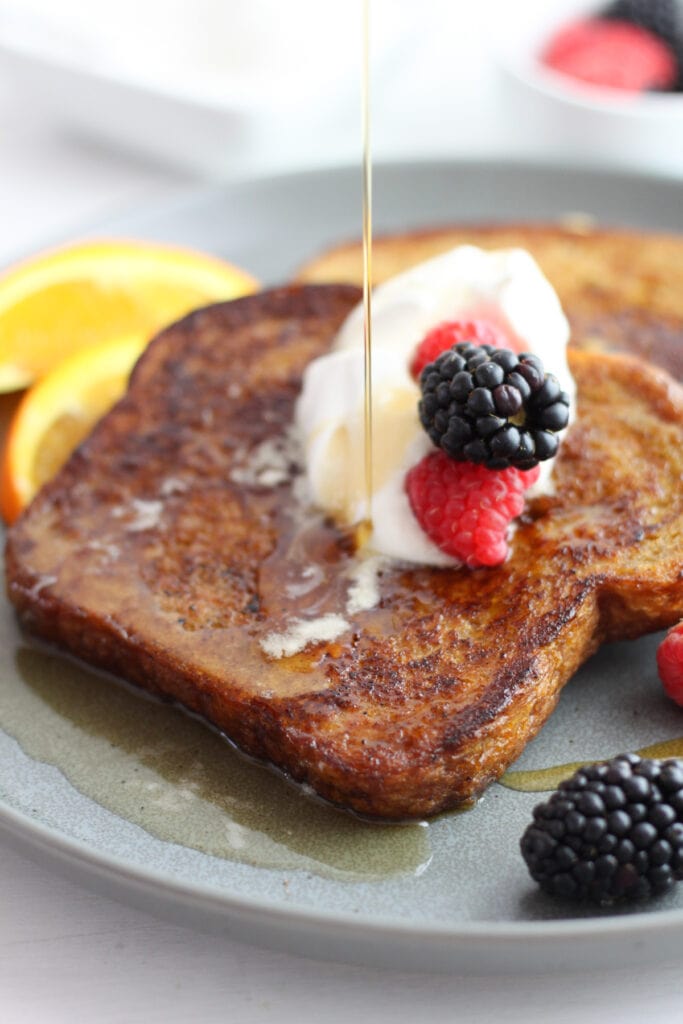 A gray plate with two slices of brown sugar french toast with whipped cream and berries and maple syrup being poured on top.