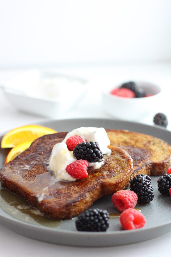 Two pieces of brown sugar French toast on a gray plate topped with whipped cream, berries, orange slices and maple syrup. In the background is a bowl with more berries.