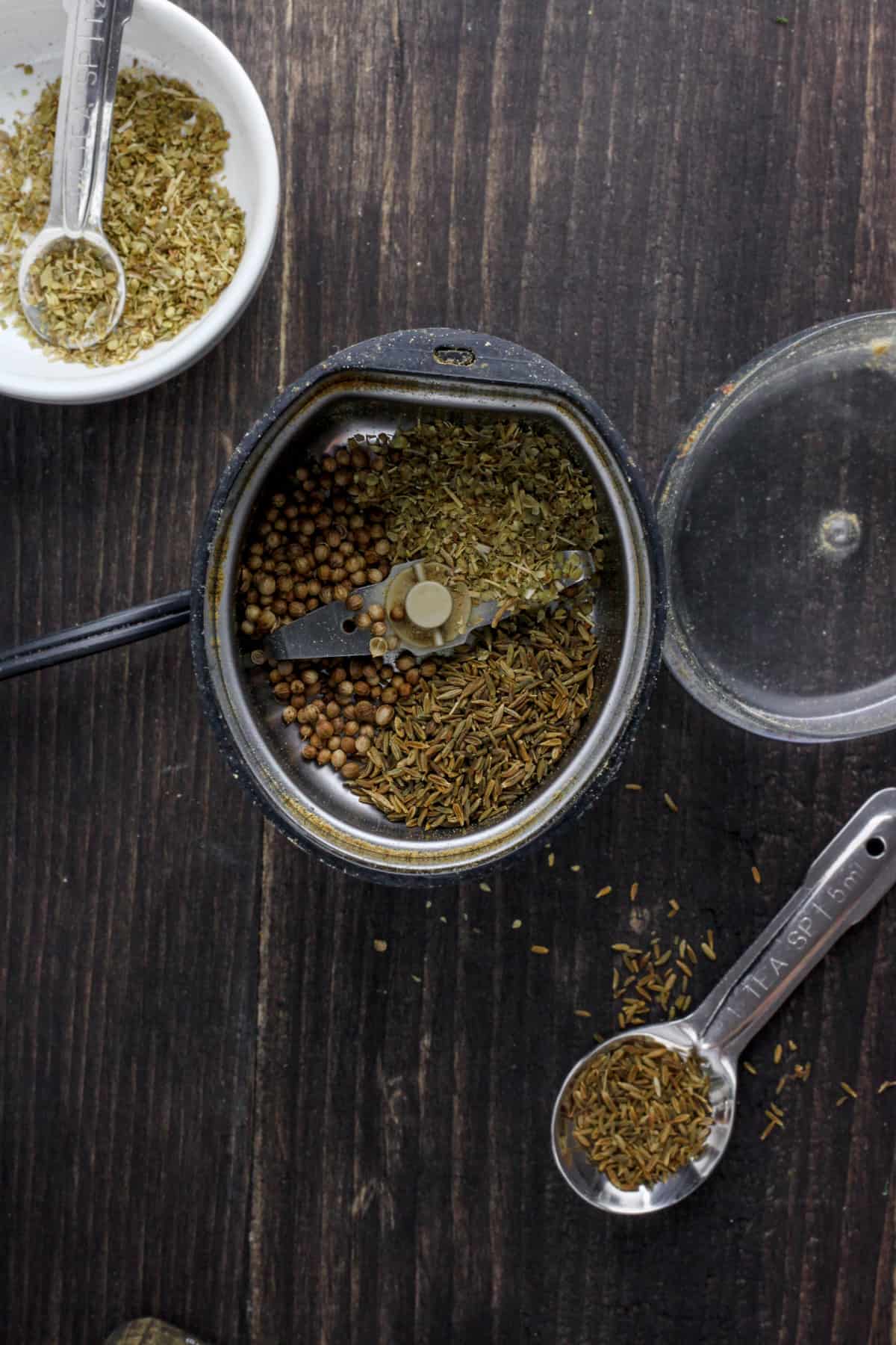 A spice grinder with whole coriander and cumin seeds and dried oregano inside.