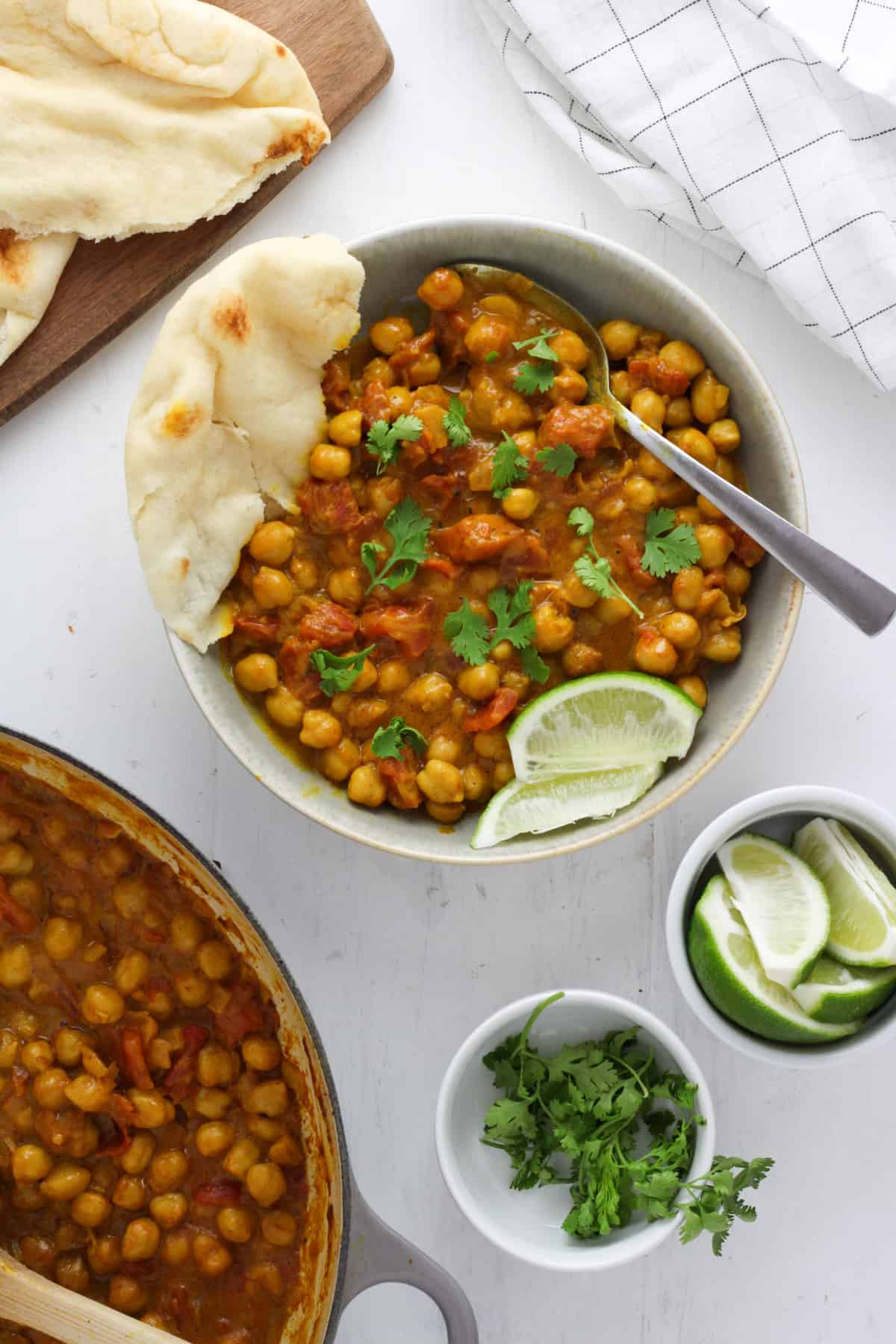 A white bowl with golden brown coconut chickpea curry, a spoon, a couple wedges of lime and a torn piece of naan. Just off center is more naan, lime wedges and more curry.