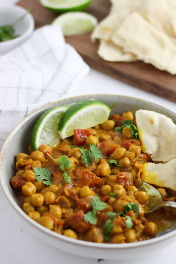 A white bowl with golden brown coconut chickpea curry, a couple wedges of limes and a torn piece of naan.