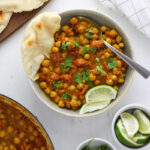A gray bowl with coconut chickpea curry topped with cilantro, lime wedges and a torn piece of naan on the side; a larger bowl of more curry is just off center along with a plate of more naan and a small bowl with limes and cilantro.