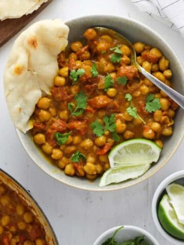 A gray bowl with coconut chickpea curry topped with cilantro, lime wedges and a torn piece of naan on the side; a larger bowl of more curry is just off center along with a plate of more naan and a small bowl with limes and cilantro.