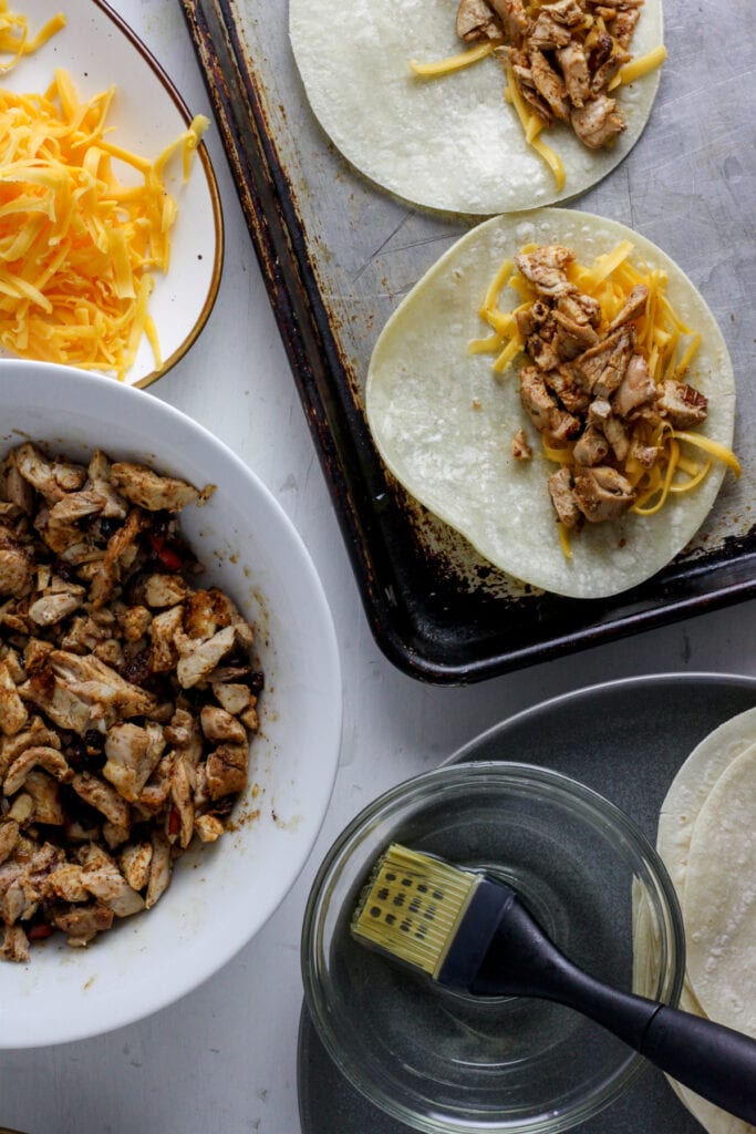 A sheet tray with corn tortillas, shredded chicken and cheese to assemble crispy chicken tacos.