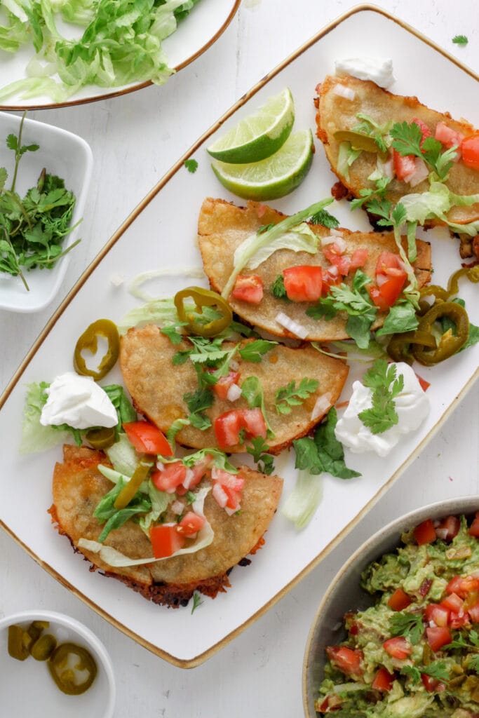 A white platter with crispy chicken tacos topped with shredded lettuce and tomatoes.