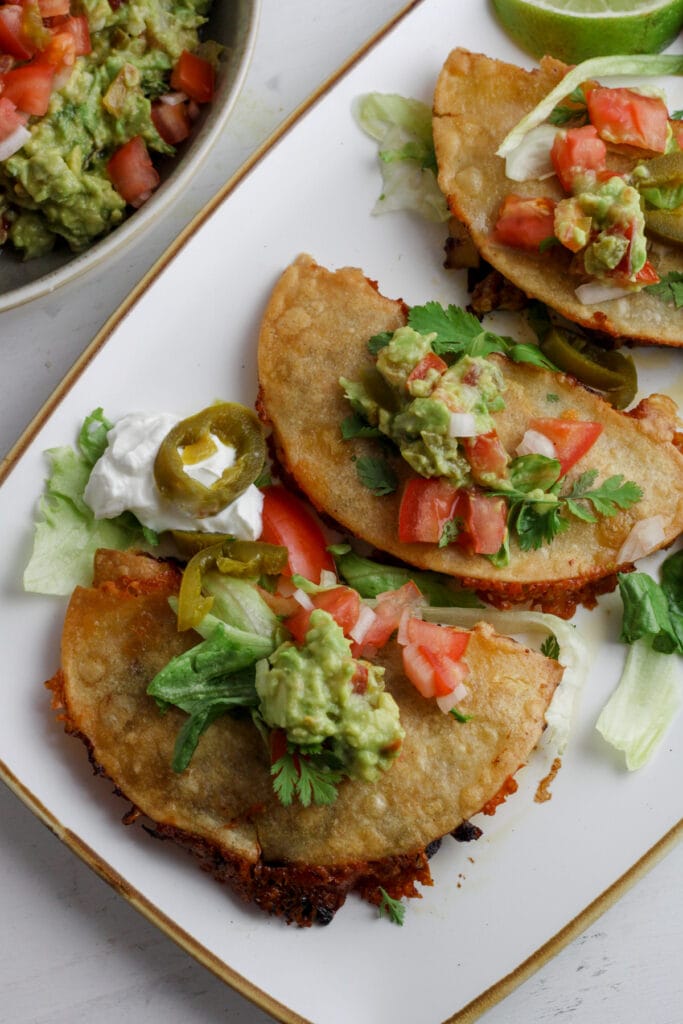 A platter of crispy chicken tacos topped with shredded lettuce and guacamole.