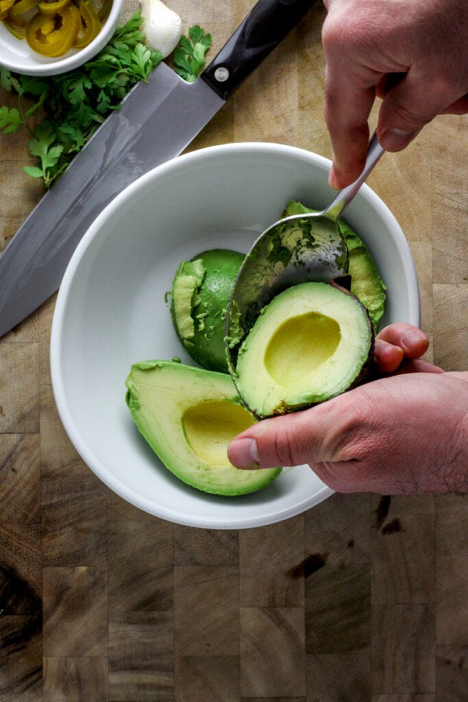 A hand using a spoon to scoop out avocado into a white bowl.