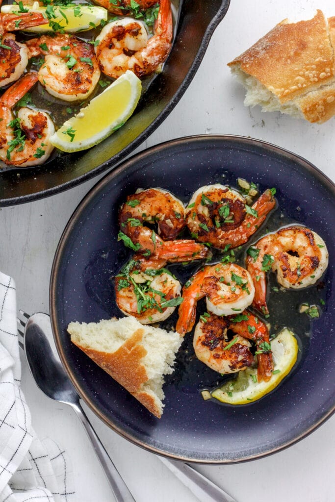 A blue plate with seared shrimp and crusty bread. off center is a skillet with more seared shrimp.