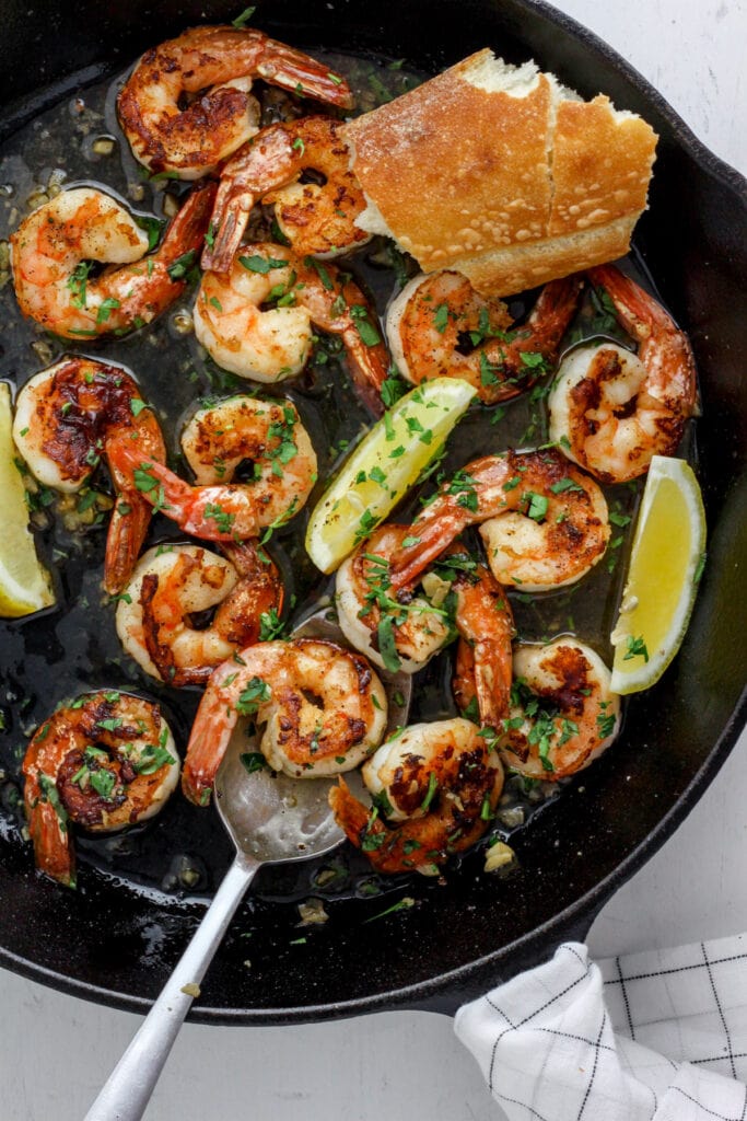 A cast iron skillet with seared shrimp, herbs, lemon wedges and crusty bread.