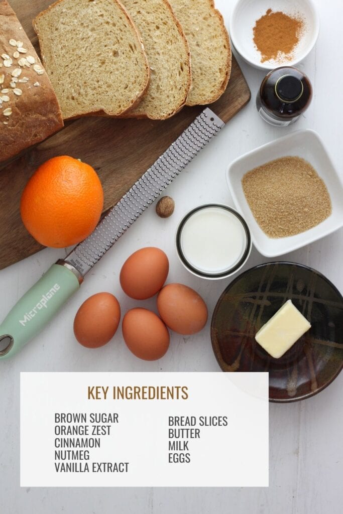 A rustic white table with a brown cutting board with bread slices, a bowl with cinnamon, a bottle of vanilla extract, another bowl with brown sugar and butter and glass of milk. There's also an orange and microplane zester, whole nutmeg and eggs.