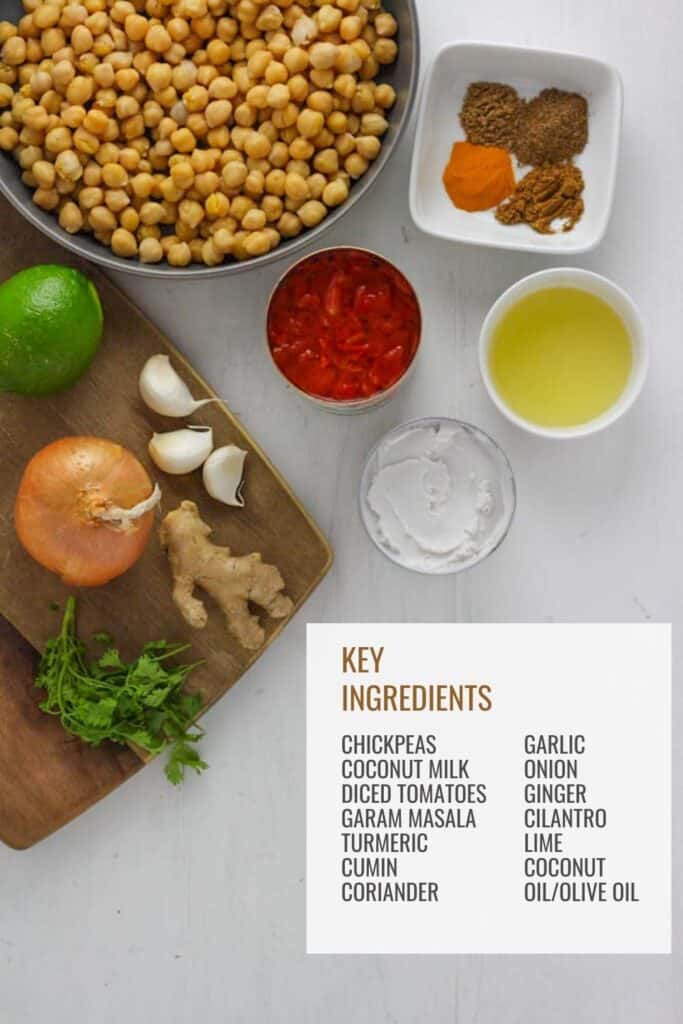 An ingredient roundup with chickpeas in a large gray bowl, a white bowl with spices like garam masala and cumin, a bowl with oil, a cutting board with onion, lime, ginger, garlic and cilantro and a can of diced tomatoes an a can of coconut milk. All of these ingredients are on a rustic white table.