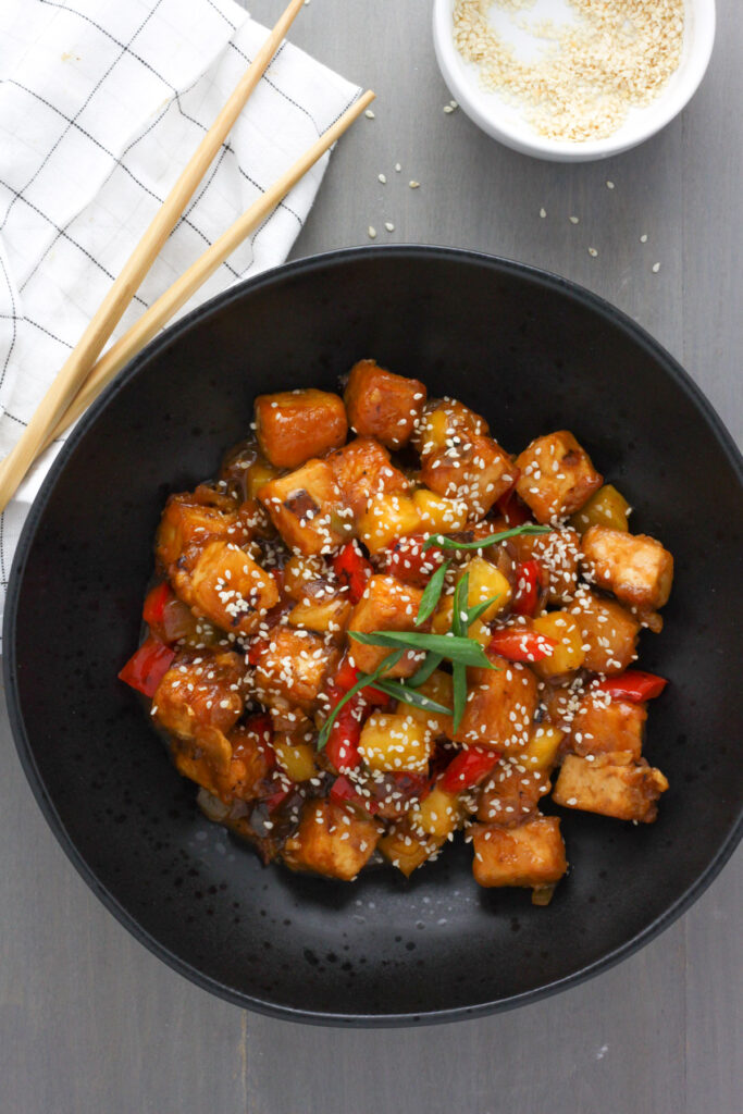 A black bowl with sweet and sour tofu topped with sesame seeds and scallions and a small white bowl with sesame seeds and a napkin just off center.