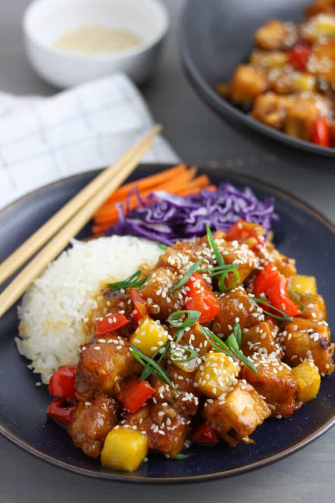 A dark blue bowl with white rice topped with sweet and sour tofu along with matchstick carrots and shredded red cabbage. There are bamboo chopsticks on the edge of the bowl and more sweet and sour tofu off center and a small bowl of sesame seeds.